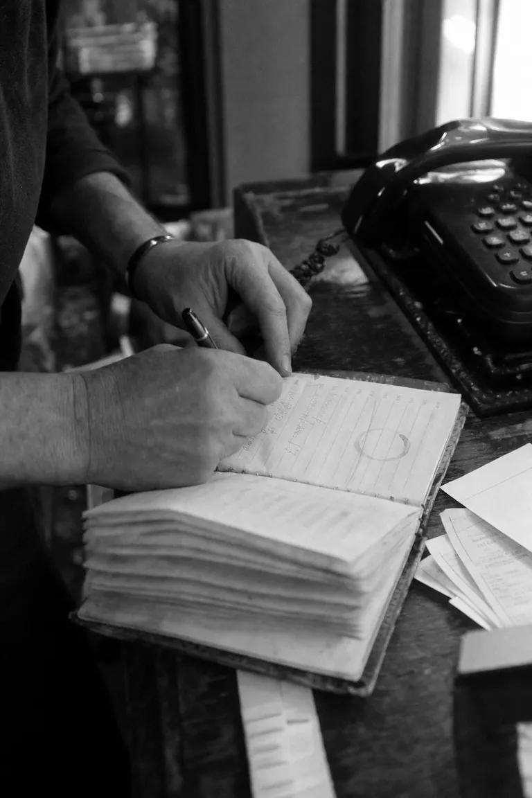 A shop owner writing in a worn paper appointment book next to a phone on the counter.